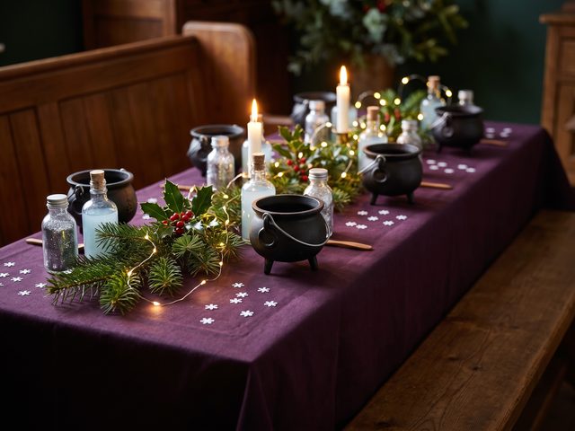 Small cast-iron cauldrons and glitter potion bottles among pine and holly by candlelight