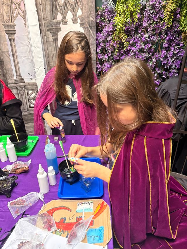 A girl in a purple cloak pouring a potion ingredient at the bench