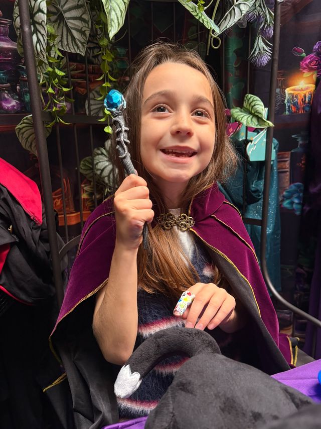 A smiling girl in a cloak holding up her wand