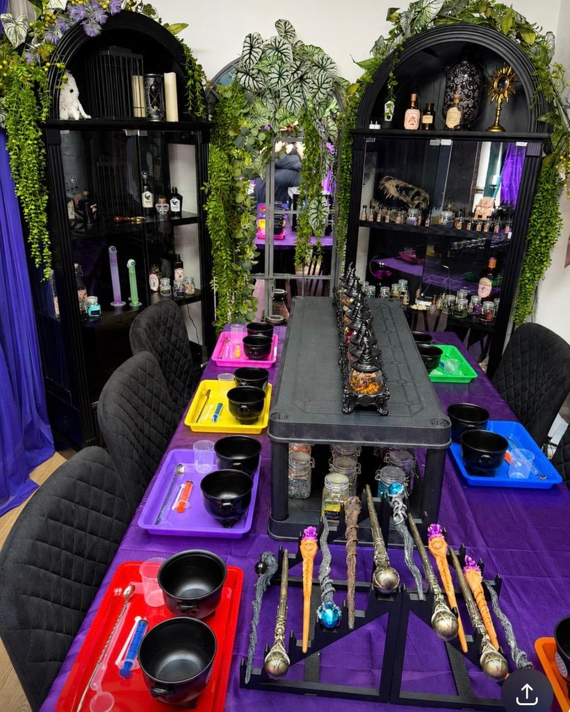 The Potion Room: cauldrons and wands laid out on a purple cloth, lit cabinets of potion bottles, trailing plants over black arched shelves