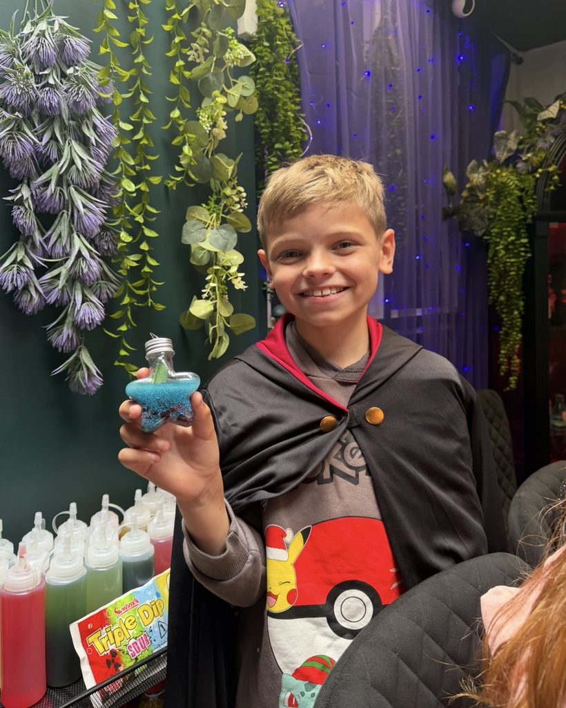 A boy in a wizard cloak grinning as he holds up his blue star-shaped potion bottle